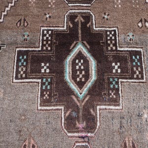 K&ouml;nnte beinhalten: Nahaufnahme eines braun-grau gemusterten Teppichs. Der Teppich weist ein zentrales geometrisches Design in Dunkelbraun auf, das wei&szlig; umrandet ist und t&uuml;rkisfarbene Akzente aufweist. Der Hintergrund ist ein helleres Graubraun mit sich wiederholenden geometrischen Formen.