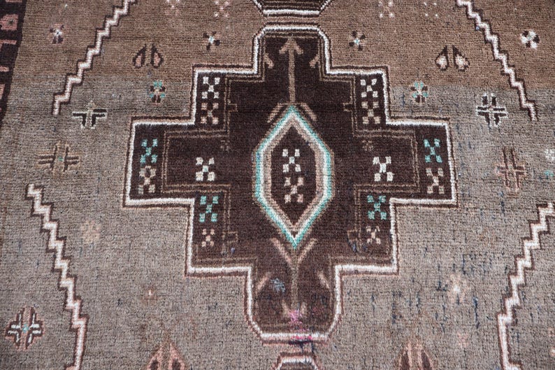 K&ouml;nnte beinhalten: Nahaufnahme eines braun-grau gemusterten Teppichs. Der Teppich weist ein zentrales geometrisches Design in Dunkelbraun auf, das wei&szlig; umrandet ist und t&uuml;rkisfarbene Akzente aufweist. Der Hintergrund ist ein helleres Graubraun mit sich wiederholenden geometrischen Formen.