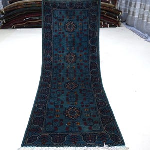 May include: Teal-colored, rectangular rug with a detailed, symmetrical pattern in dark blue and red. The rug features a central design with repeating floral motifs and a decorative border. The rug has a fringe on the bottom edge.