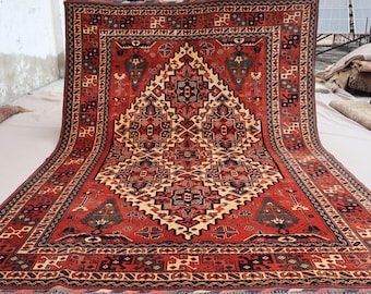 May include: A large, rectangular, hand-woven rug with a central diamond pattern in cream, black, and brown on a red background. The rug features intricate geometric designs and a detailed border. The rug is displayed on a white surface.