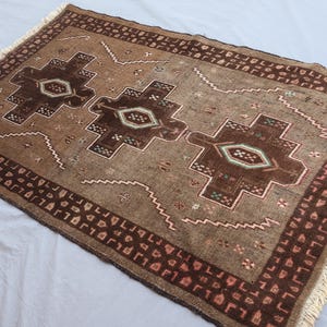 K&ouml;nnte beinhalten: Ein rechteckiger brauner Teppich mit geometrischem Muster. Der Teppich hat ein zentrales Design aus drei dunkelbraunen Formen mit helleren Akzenten, umgeben von einem Rand mit sich wiederholenden Mustern. Der Teppich hat einen Fransenrand.
