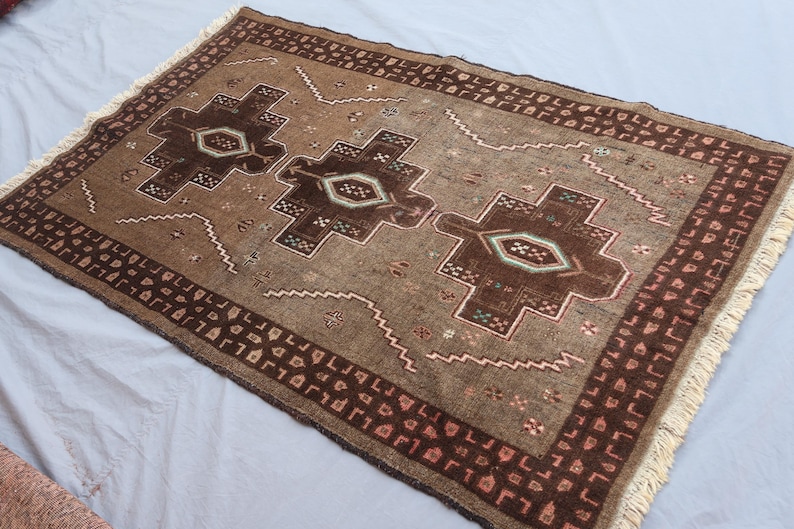 K&ouml;nnte beinhalten: Ein rechteckiger brauner Teppich mit geometrischem Muster. Der Teppich hat ein zentrales Design aus drei dunkelbraunen Formen mit helleren Akzenten, umgeben von einem Rand mit sich wiederholenden Mustern. Der Teppich hat einen Fransenrand.