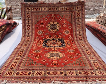 May include: A large, rectangular red rug with intricate geometric patterns in shades of beige, green, and navy blue. The rug has a central medallion design and a detailed border, with fringed edges.