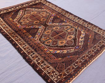 May include: A rectangular, hand-woven rug with a central geometric design in shades of brown, tan, and gold. The rug features intricate patterns and borders with accents of blue, red, and yellow. The rug's dimensions are not visible in the image.