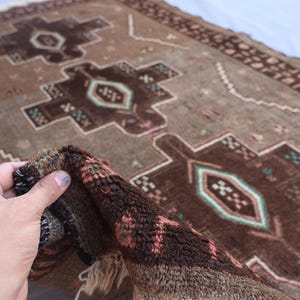 K&ouml;nnte beinhalten: Ein Vintage-Teppich in Braun- und Beiget&ouml;nen mit geometrischen Mustern. Der Teppich zeigt ein zentrales Design mit Diamanten und rechteckigen Formen in Braun, Beige und T&uuml;rkis. Der Teppich ist gefaltet und zeigt die strukturierte Unterseite.
