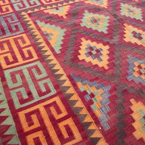 May include: A hand-woven rug with a geometric pattern. The rug features a deep red background with repeating patterns in orange, yellow, green, and blue. The border has a repeating geometric design. The rug is likely made of wool.