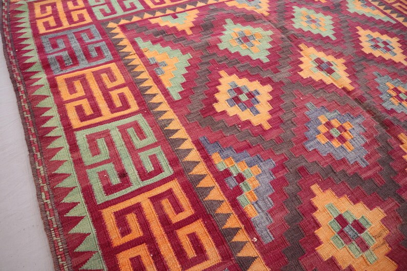 May include: A hand-woven rug with a geometric pattern. The rug features a deep red background with repeating patterns in orange, yellow, green, and blue. The border has a repeating geometric design. The rug is likely made of wool.