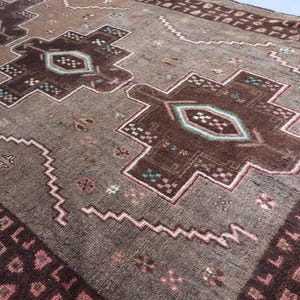 K&ouml;nnte beinhalten: Nahaufnahme eines braunen und beigen Teppichs mit einem aufwendigen geometrischen Muster. Der Teppich zeigt gro&szlig;e, stilisierte Blumenmuster in Dunkelbraun und T&uuml;rkis, eingerahmt von einem helleren beigen Rand mit sich wiederholenden Formen. Das Design umfasst Zickzacklinien und kleine florale Akzente.