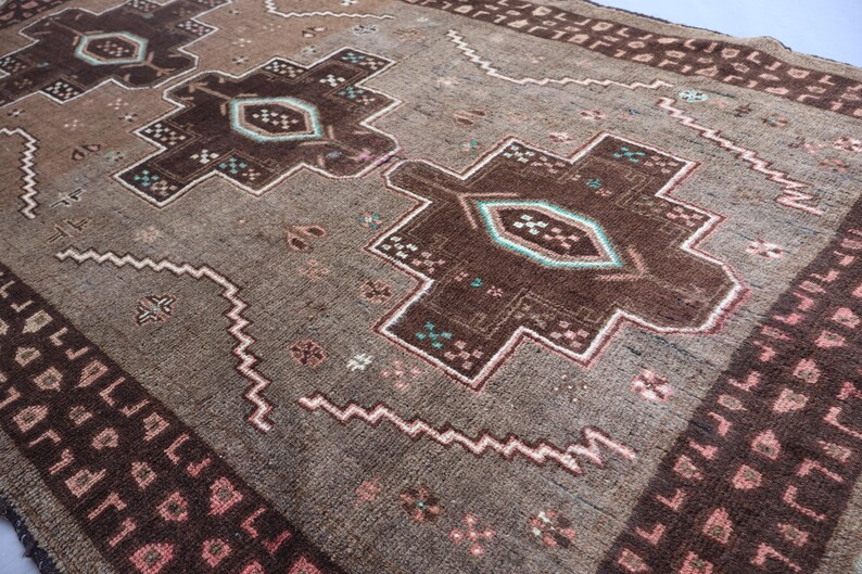 K&ouml;nnte beinhalten: Nahaufnahme eines braunen und beigen Teppichs mit einem aufwendigen geometrischen Muster. Der Teppich zeigt gro&szlig;e, stilisierte Blumenmuster in Dunkelbraun und T&uuml;rkis, eingerahmt von einem helleren beigen Rand mit sich wiederholenden Formen. Das Design umfasst Zickzacklinien und kleine florale Akzente.