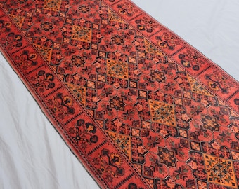 May include: A red runner rug with a complex geometric design. The rug displays a repeating pattern of black, orange, and yellow shapes on a red backdrop. The border of the rug has a detailed floral pattern.