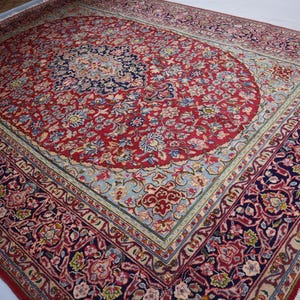 May include: A rectangular area rug featuring a central dark blue medallion surrounded by a red field filled with intricate floral and vine patterns. The border displays a complex design of flowers and leaves in shades of red, blue, and cream.