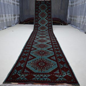 May include: A long, rectangular rug with a teal and burgundy geometric pattern. The rug features a central design of diamonds and floral motifs, with a fringe at the bottom. The rug is displayed on a white surface.