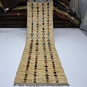 Puede incluir: Una alfombra de pasillo larga y rectangular con una base de color crema y un tejido horizontal texturizado. La alfombra está decorada con tres filas verticales de pequeños patrones en forma de rombo multicolor en tonos rojo, azul, verde y amarillo.