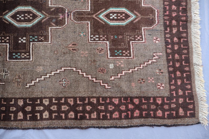 K&ouml;nnte beinhalten: Nahaufnahme eines Teils eines Vintage-Teppichs mit geometrischem Muster. Der Teppich hat eine braune und graue Farbpalette mit Akzenten in T&uuml;rkis und Rosa. Das Design umfasst sich wiederholende Formen und einen Fransenrand.