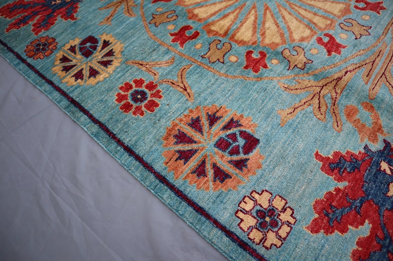 May include: A teal blue rug with a large, intricate, circular pattern in the center. The pattern is made up of beige, orange, red, and purple colors. The rug has a border of smaller floral patterns in the same colors.