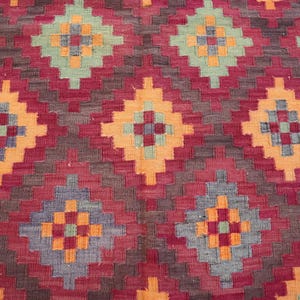 May include: Close-up of a woven rug with a geometric pattern. The rug features a repeating diamond design in shades of red, orange, blue, and green. The background is a deep red, with the diamond shapes outlined in a lighter orange.