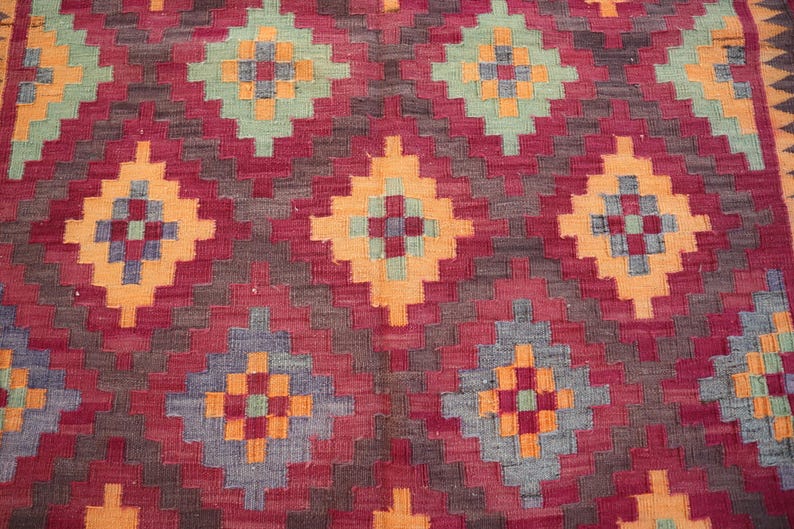 May include: Close-up of a woven rug with a geometric pattern. The rug features a repeating diamond design in shades of red, orange, blue, and green. The background is a deep red, with the diamond shapes outlined in a lighter orange.