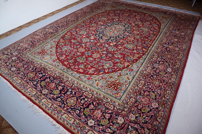 May include: A large rectangular area rug featuring a central red oval medallion filled with intricate floral patterns in blue, cream, and gold. The surrounding field is light blue, framed by a wide dark blue border with dense floral motifs.