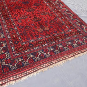 May include: A rectangular, hand-woven rug with a deep red field and intricate geometric patterns in navy blue, brown, and cream. The rug features a detailed border and a fringed edge, showcasing traditional craftsmanship.