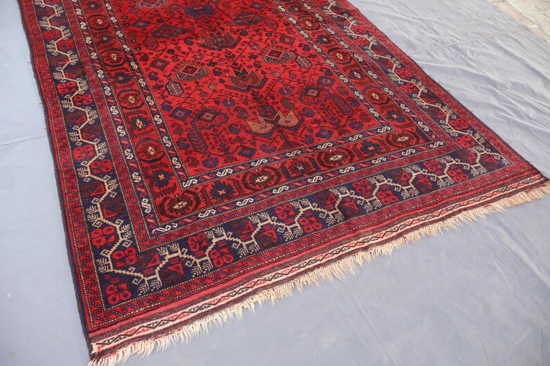 May include: A rectangular, hand-woven rug with a deep red field and intricate geometric patterns in navy blue, brown, and cream. The rug features a detailed border and a fringed edge, showcasing traditional craftsmanship.