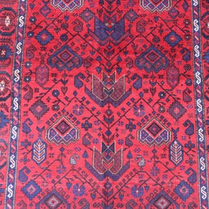 May include: A red and blue patterned rug with a central design of stylized floral and geometric shapes. The rug features a repeating pattern with a border of alternating shapes and colors. The rug is likely made of wool.