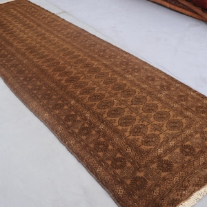 Peut inclure: Un long tapis rectangulaire marron avec un motif floral complexe. Le tapis présente un motif central de motifs circulaires répétitifs et une bordure détaillée. La frange du tapis est de couleur beige clair.