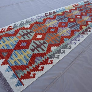 May include: A white rug with a colourful geometric pattern in shades of red, orange, blue, green, and yellow. The rug has a fringe border.
