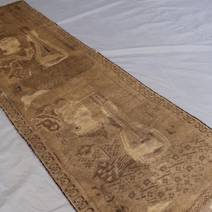 May include: A rectangular, brown rug with a detailed design featuring musical instruments and figures. The rug has a patterned border and fringe on one end. The design includes a lute and other musical elements.