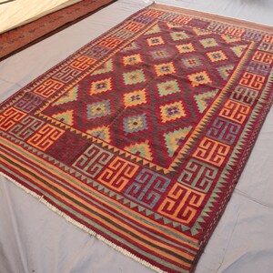 May include: A rectangular, hand-woven rug with a geometric pattern. The rug features a deep red central field with diamond shapes in shades of yellow, green, and blue. The border has a repeating geometric design in red, orange, and blue.