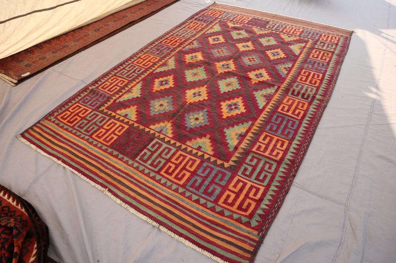 May include: A rectangular, hand-woven rug with a geometric pattern. The rug features a deep red central field with diamond shapes in shades of yellow, green, and blue. The border has a repeating geometric design in red, orange, and blue.