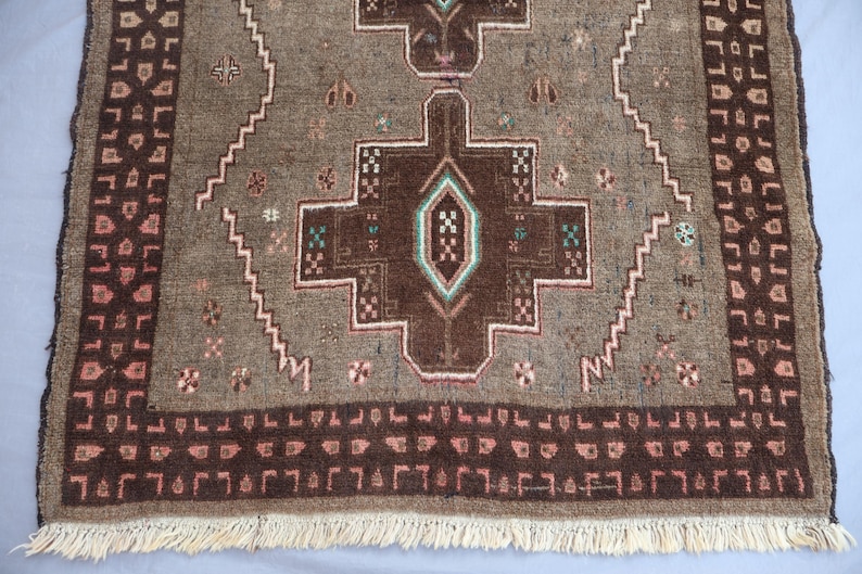 K&ouml;nnte beinhalten: Ein rechteckiger, handgewebter Teppich mit einem zentralen geometrischen Design in Braun-, Grau- und T&uuml;rkist&ouml;nen. Der Teppich hat einen detaillierten Rand mit sich wiederholenden Mustern und eine Fransen am unteren Rand. Die Abmessungen des Teppichs sind nicht sichtbar.