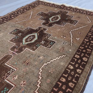 K&ouml;nnte beinhalten: Ein rechteckiger brauner Teppich mit einem zentralen geometrischen Design in dunklerem Braun und T&uuml;rkis. Der Teppich hat einen Rand mit einem sich wiederholenden Muster und Fransen an einem Ende. Der Teppich ist wahrscheinlich aus Wolle oder einem &auml;hnlichen Material.