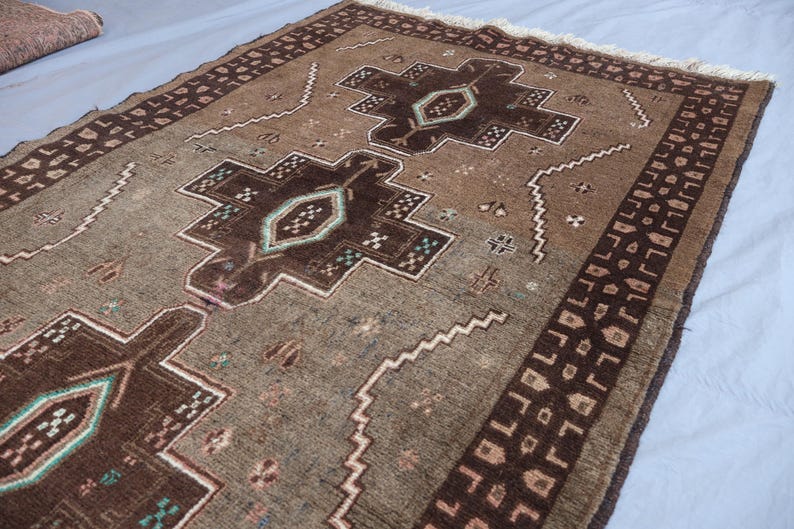 K&ouml;nnte beinhalten: Ein rechteckiger brauner Teppich mit einem zentralen geometrischen Design in dunklerem Braun und T&uuml;rkis. Der Teppich hat einen Rand mit einem sich wiederholenden Muster und Fransen an einem Ende. Der Teppich ist wahrscheinlich aus Wolle oder einem &auml;hnlichen Material.