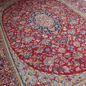 May include: A rectangular area rug featuring a deep red central field with an intricate floral pattern in shades of blue, cream, and pink. The rug has a prominent dark blue central medallion and a wide, detailed border with complex floral motifs.