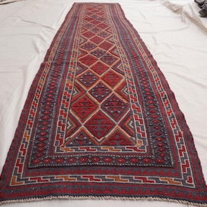 May include: A long, narrow rug with a traditional geometric pattern in shades of red, blue, and cream. The rug has a fringe border and is made of wool.