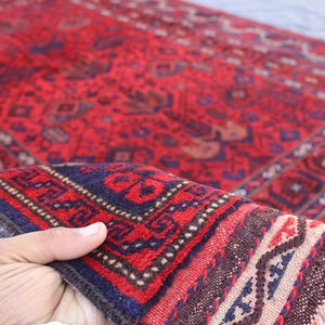 May include: A hand-woven red rug with intricate geometric patterns in navy blue, brown, and cream. The rug features a detailed border and fringed edges, showcasing traditional craftsmanship. The rug is a decorative floor covering.
