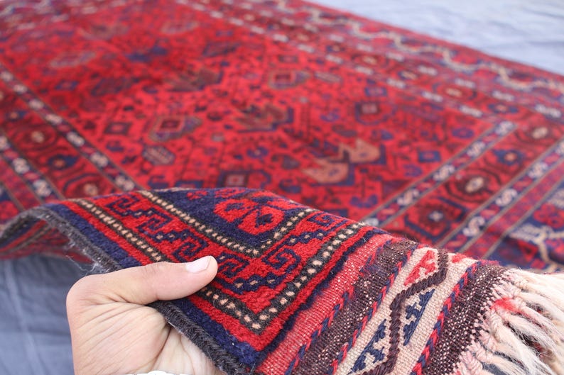 May include: A hand-woven red rug with intricate geometric patterns in navy blue, brown, and cream. The rug features a detailed border and fringed edges, showcasing traditional craftsmanship. The rug is a decorative floor covering.