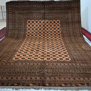 May include: A large rectangular area rug featuring a central field of light orange with a repeating dark brown circular geometric pattern. The rug is framed by multiple wide borders in shades of dark brown with intricate linear and geometric motifs.