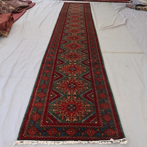 Puede incluir: Una alfombra larga y rectangular con un patrón detallado. La alfombra presenta un diseño central en rojo y verde, con intrincados motivos florales y geométricos. Los bordes son de un rojo más oscuro con detalles azules, y la alfombra tiene una franja blanca.