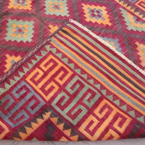 May include: A detailed shot of a red, geometric patterned rug. The rug features repeating square and rectangular designs in shades of red, orange, green, and gray. The edge of the rug is folded over, revealing a striped border.