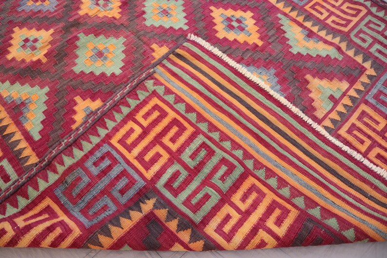 May include: A detailed shot of a red, geometric patterned rug. The rug features repeating square and rectangular designs in shades of red, orange, green, and gray. The edge of the rug is folded over, revealing a striped border.