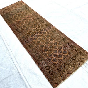May include: A long rectangular runner rug featuring a repeating geometric pattern of octagonal shapes in shades of brown, tan, and muted orange. The rug has a detailed border with intricate diamond and linear motifs and short fringe at both ends.