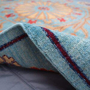 May include: Close-up of a blue rug with a red and cream floral pattern. The rug is folded over, showing the underside with a red stripe.