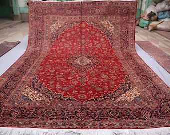 May include: A large rectangular area rug featuring a vibrant red field with a central dark blue medallion and intricate floral patterns in shades of cream, navy, and gold. The rug has a wide, detailed border and white fringe along the bottom edge.