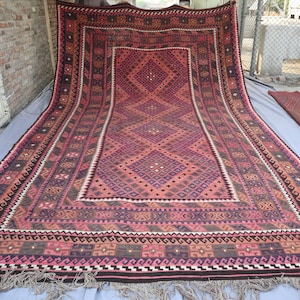 May include: A large, rectangular rug with a geometric pattern in shades of red, pink, purple, and brown. The rug features a central diamond design and a detailed border with fringe along the edges. The rug is displayed outdoors.