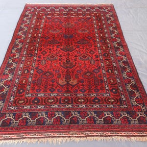 May include: A rectangular red rug with an intricate geometric pattern. The rug features a central field of red with a detailed border in navy blue, brown, and cream. The rug has a fringed edge.