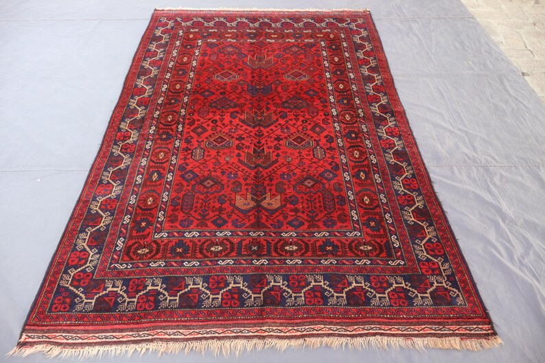 May include: A rectangular red rug with an intricate geometric pattern. The rug features a central field of red with a detailed border in navy blue, brown, and cream. The rug has a fringed edge.