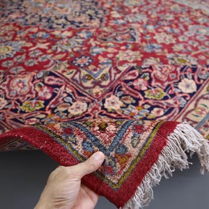 May include: A red area rug featuring an intricate floral pattern in shades of blue, cream, and gold. The rug has a dense pile, a dark blue border with detailed botanical motifs, and white fringe along the edge where the corner is folded over.