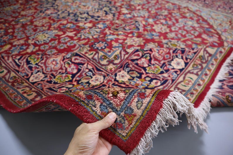 May include: A red area rug featuring an intricate floral pattern in shades of blue, cream, and gold. The rug has a dense pile, a dark blue border with detailed botanical motifs, and white fringe along the edge where the corner is folded over.