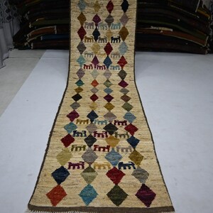 May include: A long, narrow runner rug with a cream-colored base featuring a repeating pattern of multicolored diamonds and small, stylized animal figures. The rug has a dark brown border and a fringed edge at the bottom, showcasing a variety of hues.